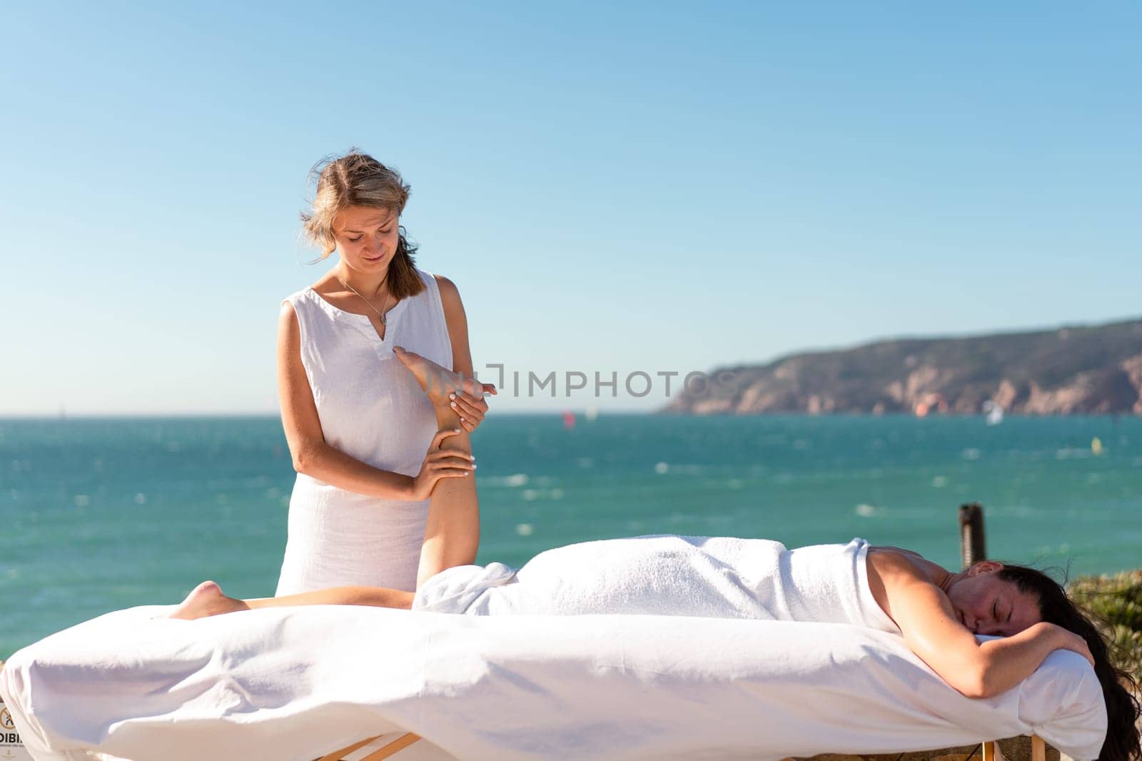 Therapist massaging woman's leg on table by sea against clear blue sky during sunny day. Masseuse is pampering female client during summertime.