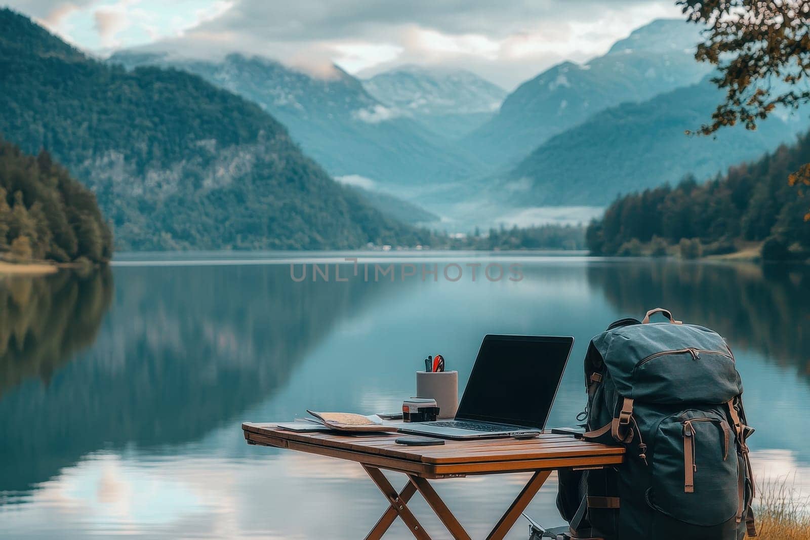 A picturesque lakeside workspace featuring a laptop and backpack, surrounded by majestic mountains, offering a perfect blend of nature and productivity.