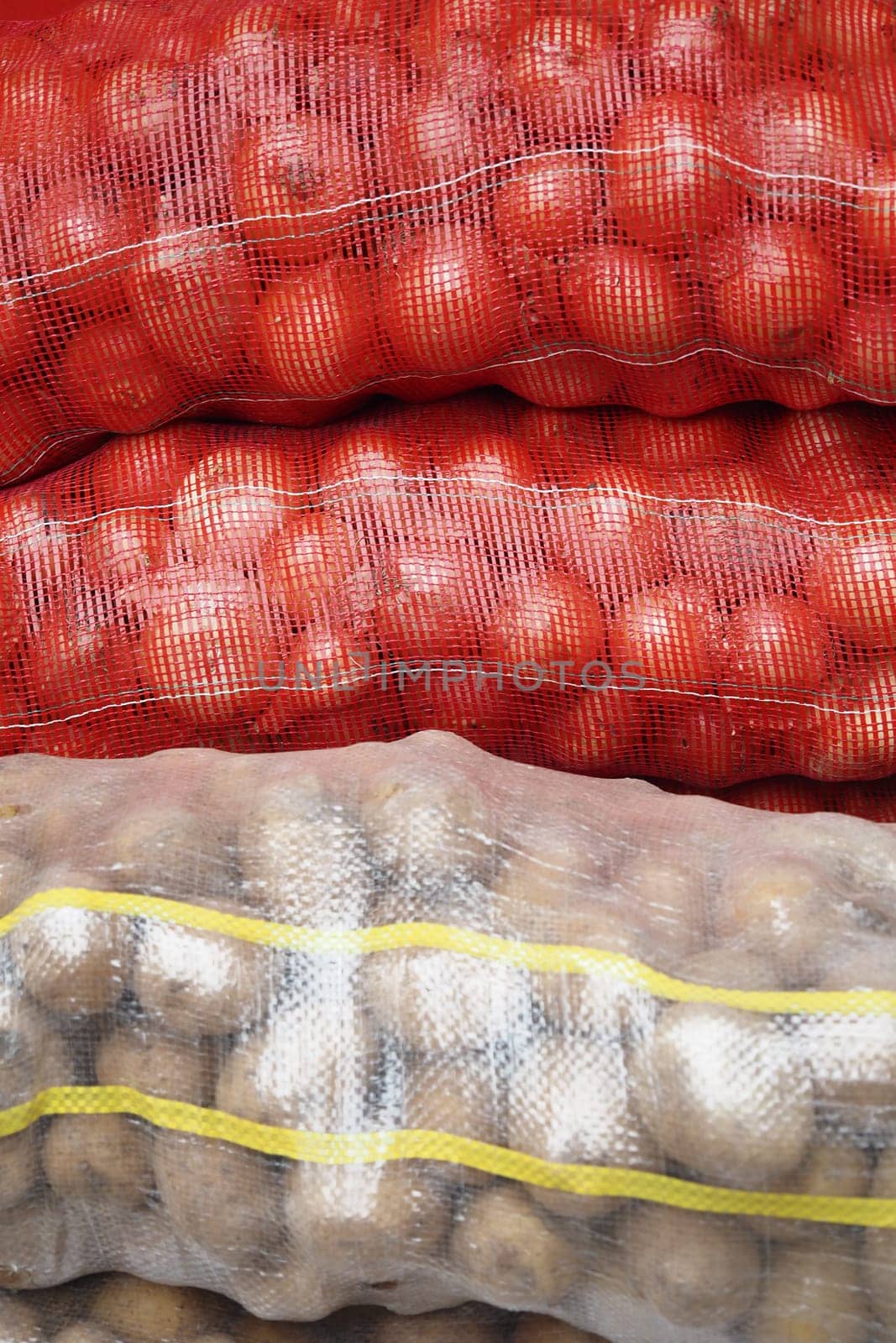 A variety of fresh bags filled with red onions, potatoes, and assorted vegetables are stacked