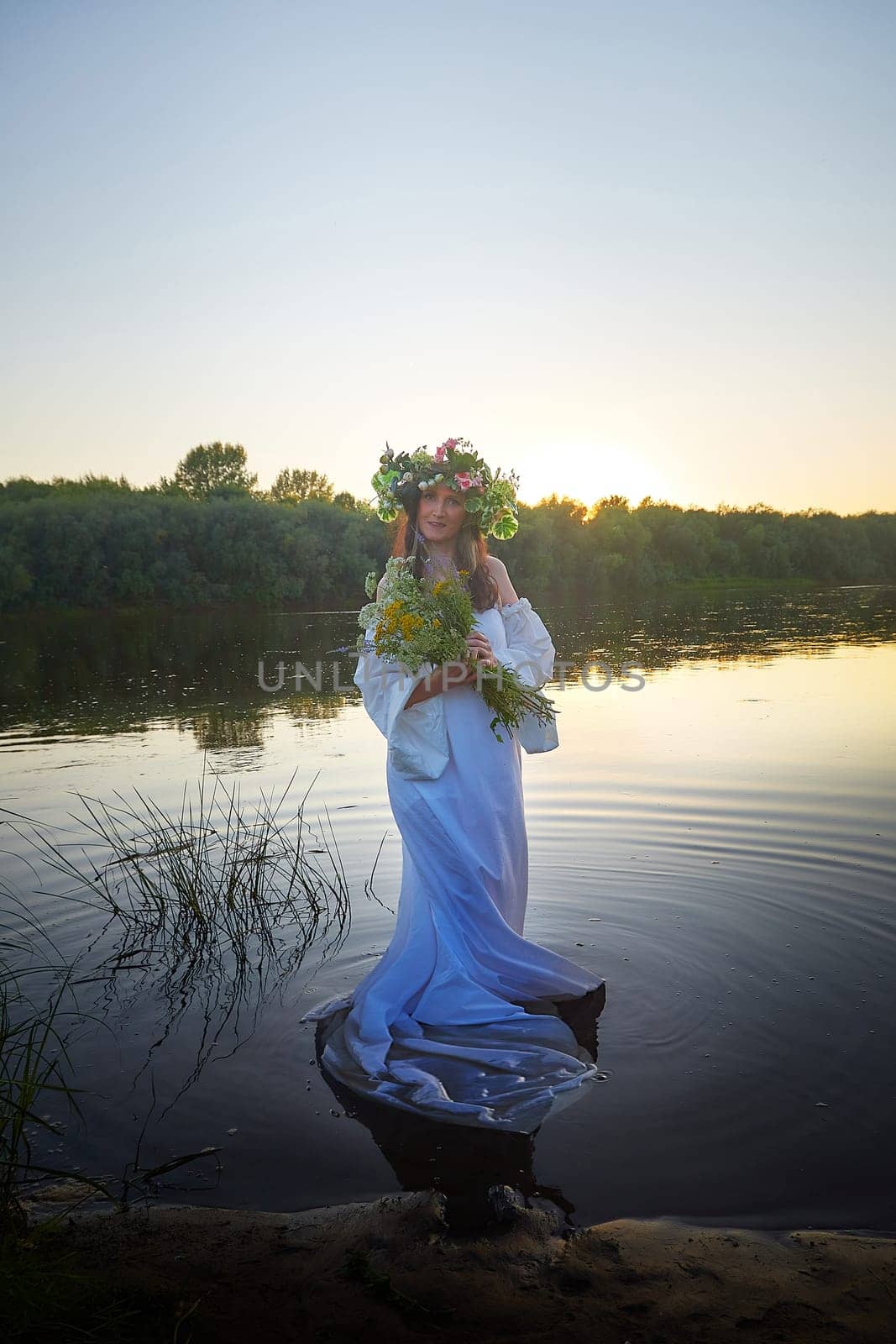 Slavic plump chubby girl in long white dress on the feast of Ivan Kupala with flowers and water in a river or lake on summer evening