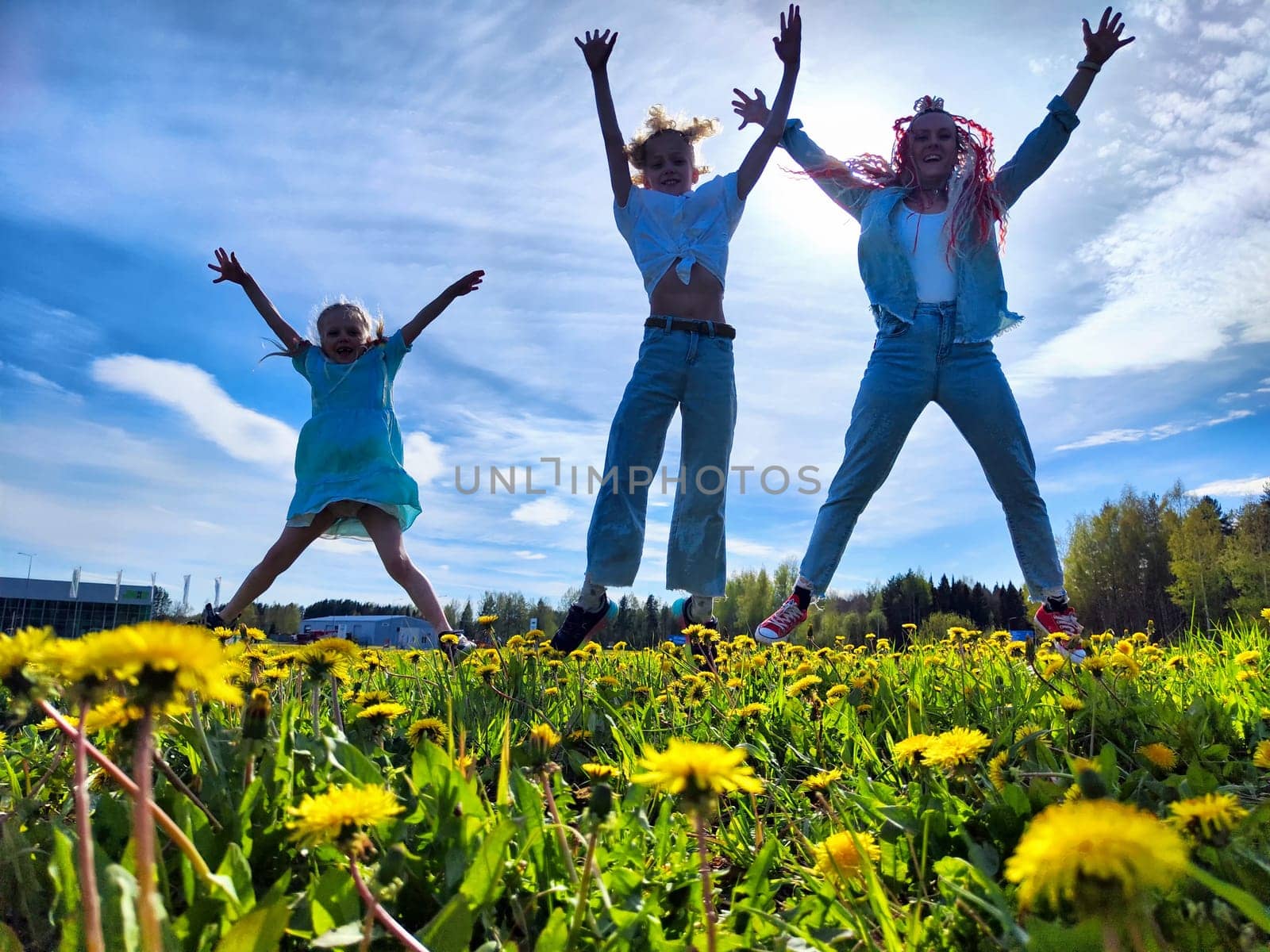 Children are leaping happily in a grassy field filled with dandelions, enjoying a beautiful spring day with clear skies.