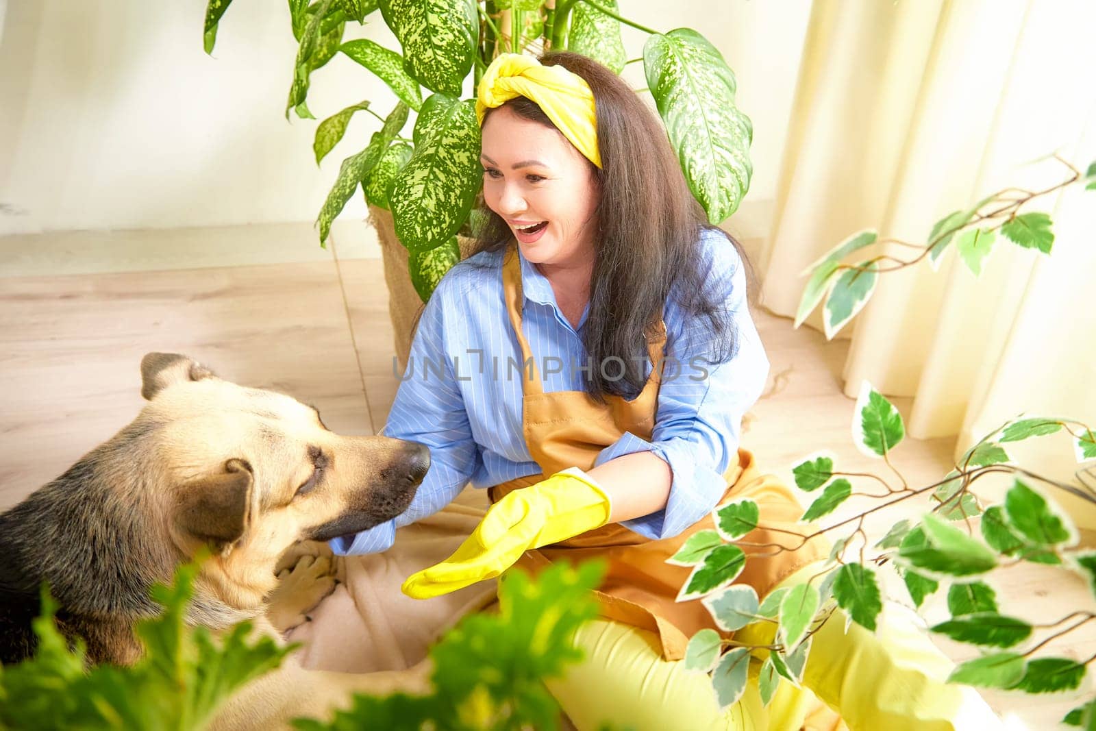 The gardener smiles as she plays with a dog among vibrant houseplants indoors.