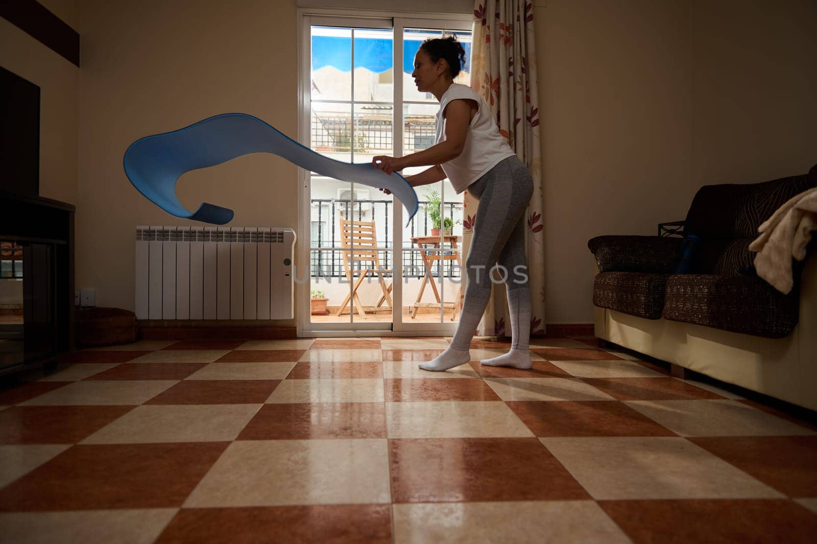 A woman engages in a stretching routine using a blue fitness band in her living room. Natural light fills the space, creating a calming environment for a focused workout session.