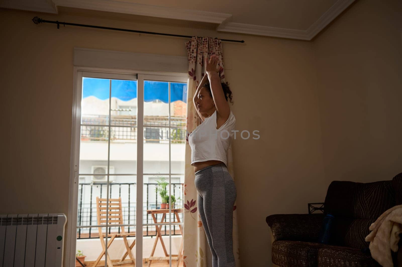 A woman performs a yoga stretch indoors with sunlight streaming through a window. The serene atmosphere enhances focus and mindfulness, reflecting a peaceful start to the day.
