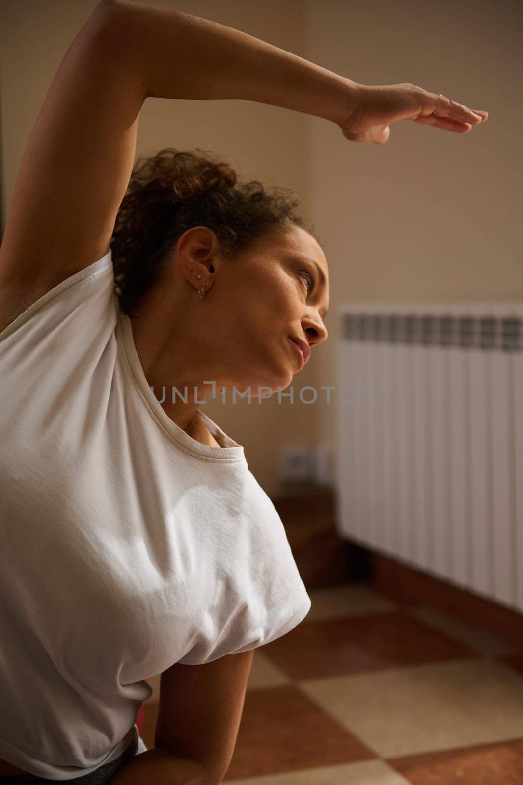 A woman in a white t-shirt practices yoga indoors, embodying calm and focus. The late afternoon light adds warmth to the serene atmosphere, highlighting her pose and determination.