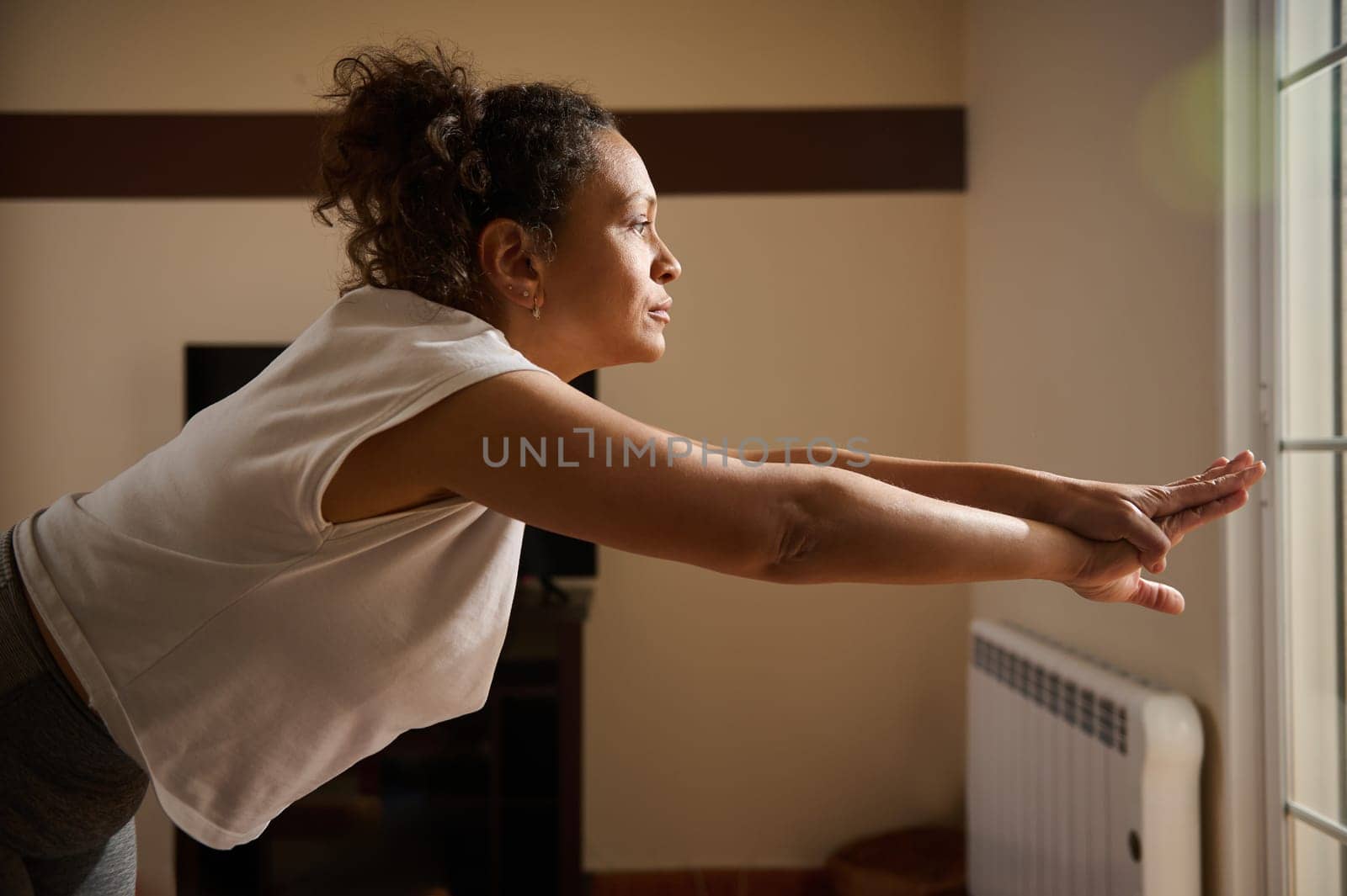 A woman is practicing yoga indoors, stretching forward with focus and calmness. The natural light highlights her peaceful expression, embodying balance and mindfulness.