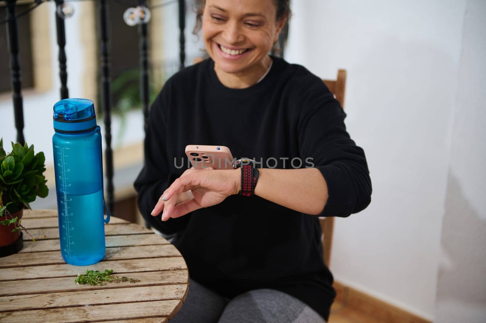A smiling woman checks her smart watch while holding a smartphone. She sits beside a blue water bottle on a wooden table, creating a relaxed atmosphere. Perfect for tech and lifestyle themes.