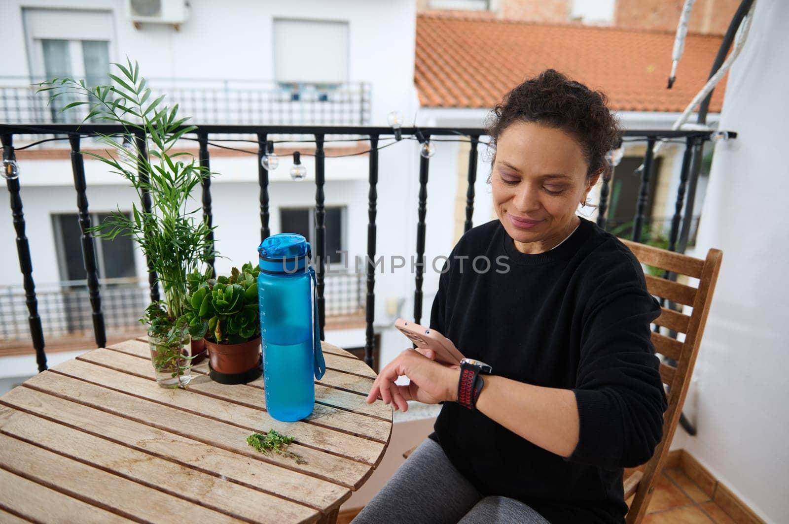 A woman sitting on a balcony checks her smartwatch, surrounded by green plants and a blue water bottle. The scene is calm and serene, highlighting relaxation and mindfulness.