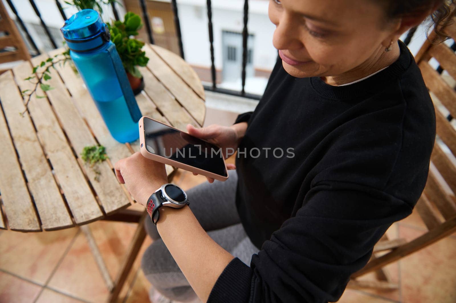A woman sits on a balcony, checking notifications on her smartwatch while holding a smartphone. A water bottle and plant are on a nearby table, reflecting a relaxed outdoor setting.