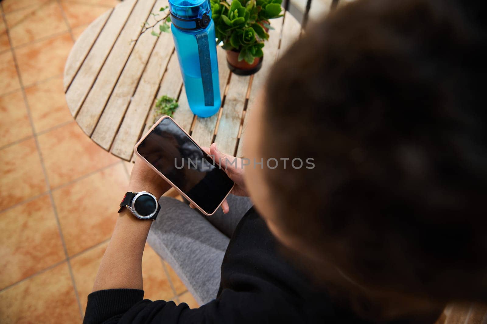 A person is seated at a wooden table, viewing a smartphone. They wear a smartwatch and have a water bottle nearby, suggesting leisure or fitness. The setting is relaxed and casual.