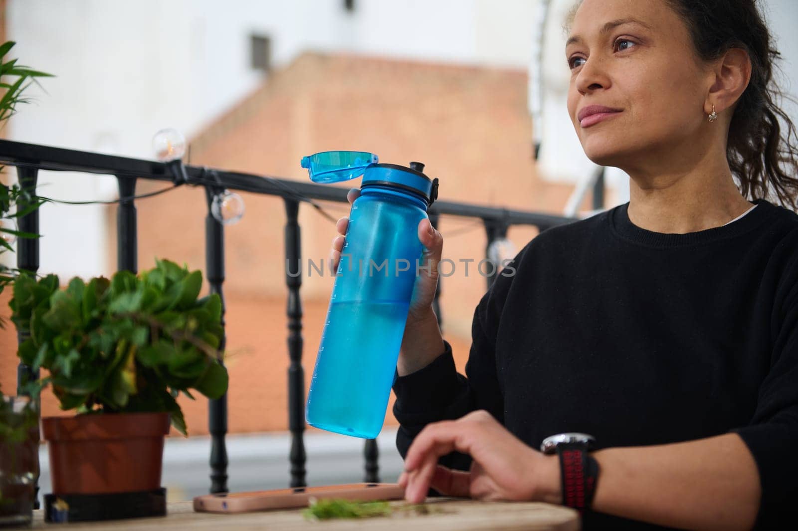 A woman sits on a balcony, holding a blue water bottle, surrounded by plants. She appears relaxed in the sunlight, symbolizing hydration and tranquility in an outdoor setting.