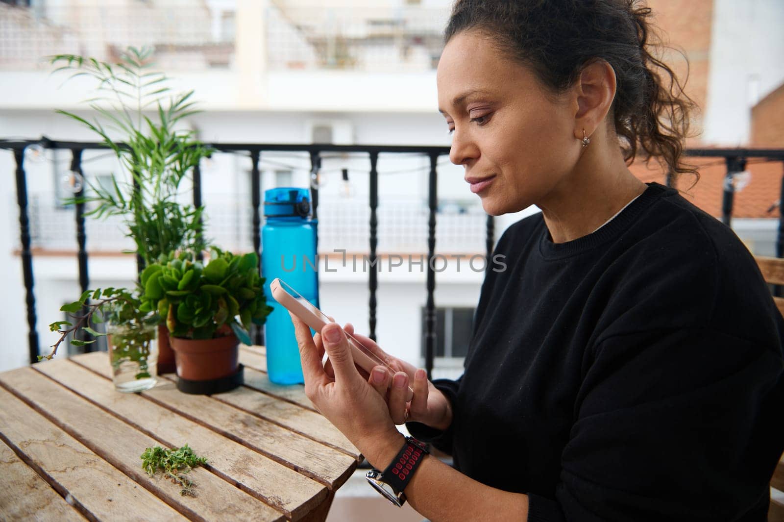 A woman sits on a balcony table texting on her smartphone. Surrounded by greenery and plants, she enjoys a tranquil moment in an urban setting, with a water bottle in the background.