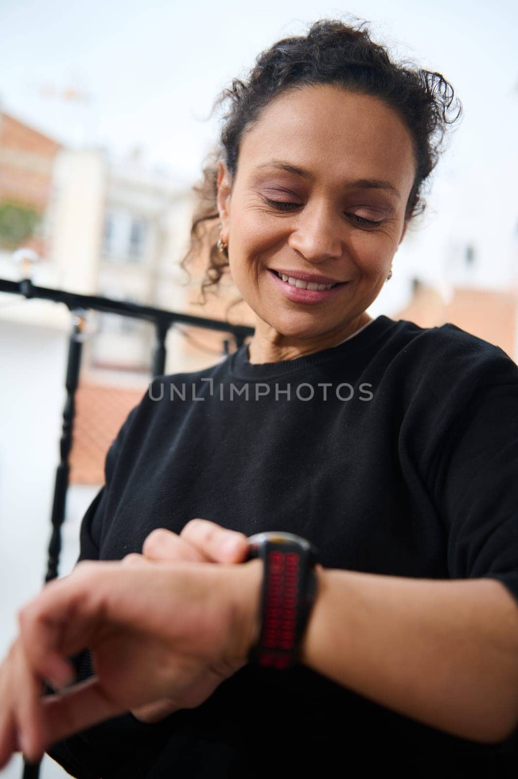 A cheerful woman looks at her smartwatch while enjoying an outdoor setting. She expresses a sense of accomplishment and happiness, perhaps after a workout or successful activity.