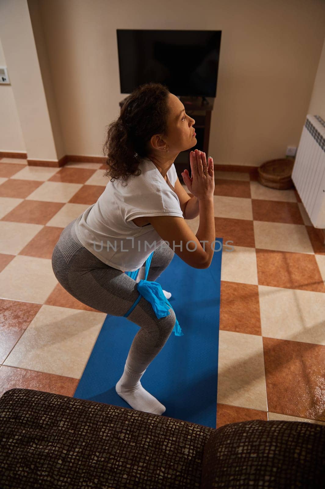 A woman performs a yoga pose on a blue mat in a cozy living room setting. The indoor environment suggests comfort and focus, highlighting relaxation and fitness in a personal space.