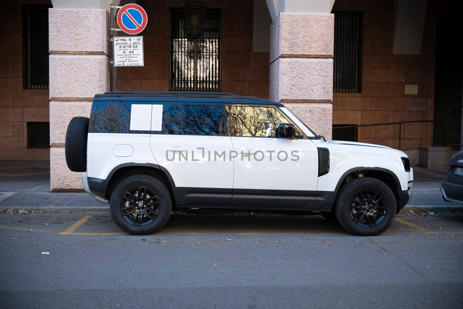Cremona, Lombardy, Italy - October 27th 2024 Side view of a white land rover defender 110 p400e suv parked on the street next to a no parking traffic sign