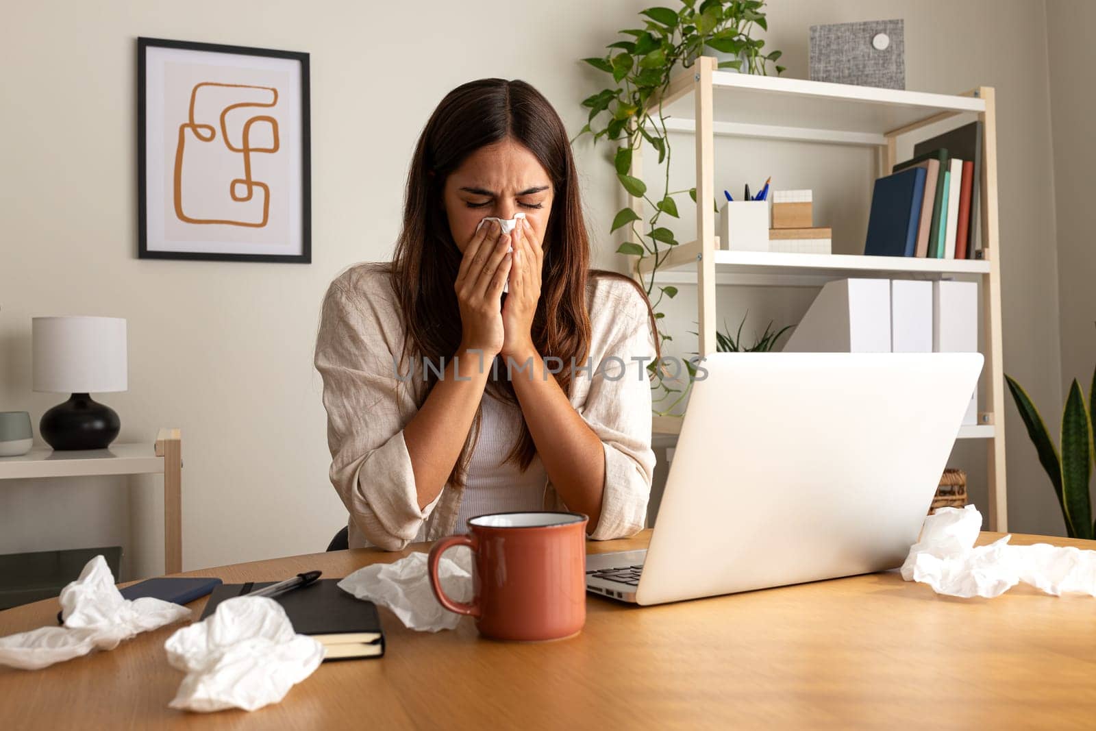 Sick woman working from home office. Caucasian female blowing nose with tissue while working with laptop. by Hoverstock