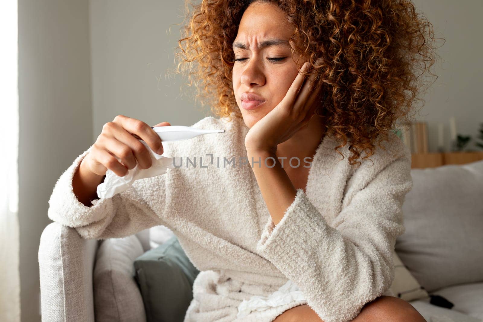 Sick woman checking temperature with thermometer, sitting on the sofa feeling unwell. Sick woman at home with flu virus having fever. Sickness concept.