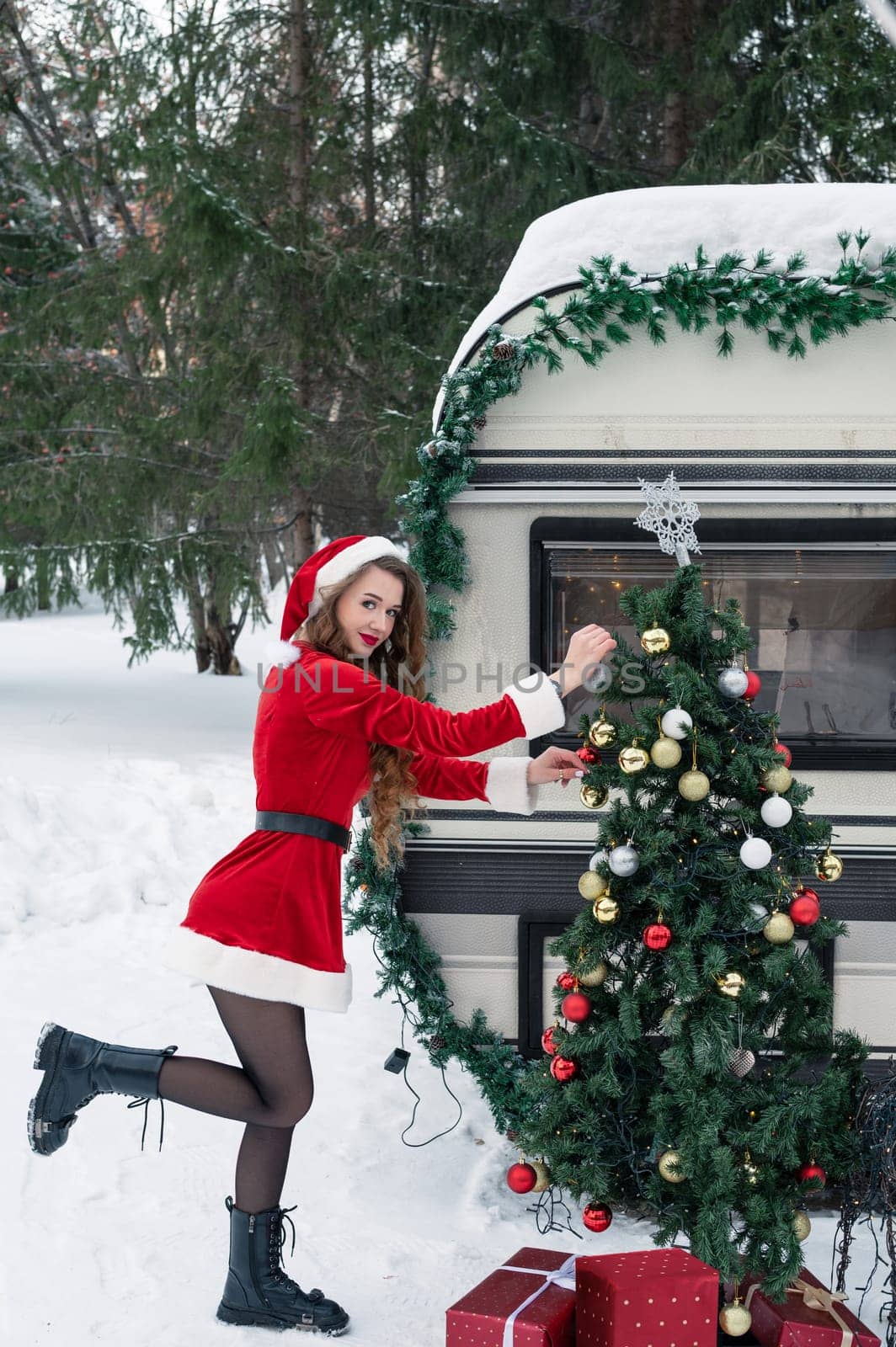 Young woman in santa costume decorates the Christmas tree at winter campsite getting ready for the new year. New year celebration concept