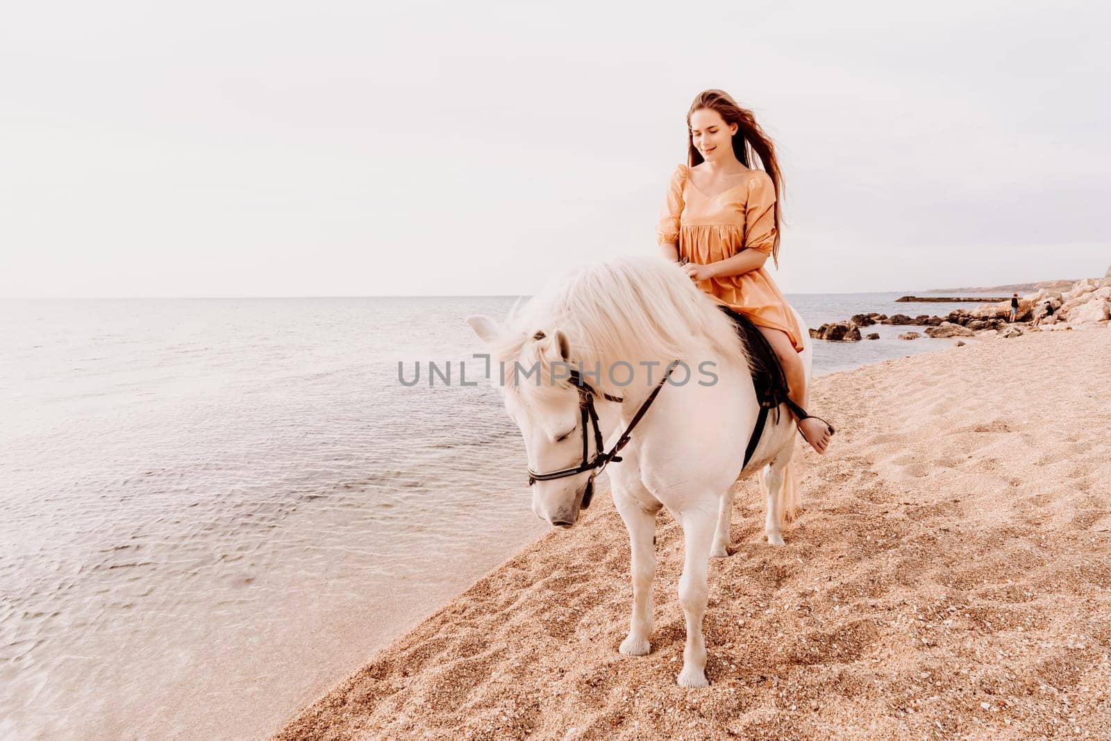 A woman in a dress stands next to a white horse on a beach, with the blue sky and sea in the background