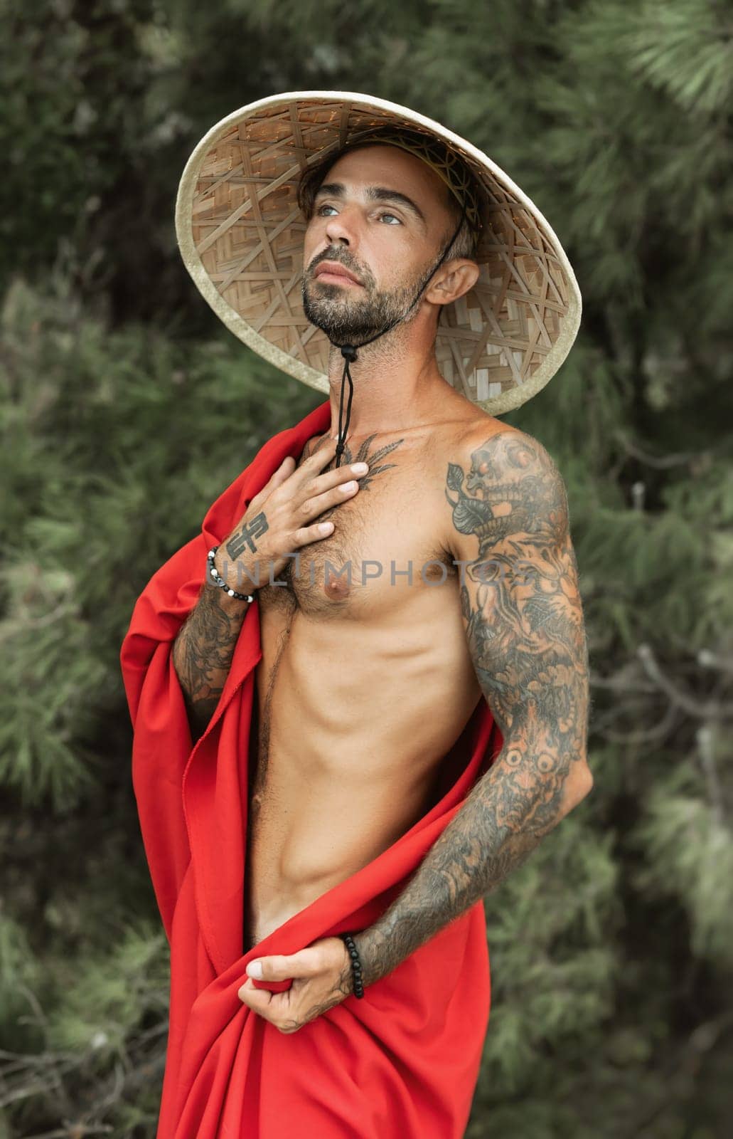 the sexy male monk in a red cape and asian hat in tattoos in nature