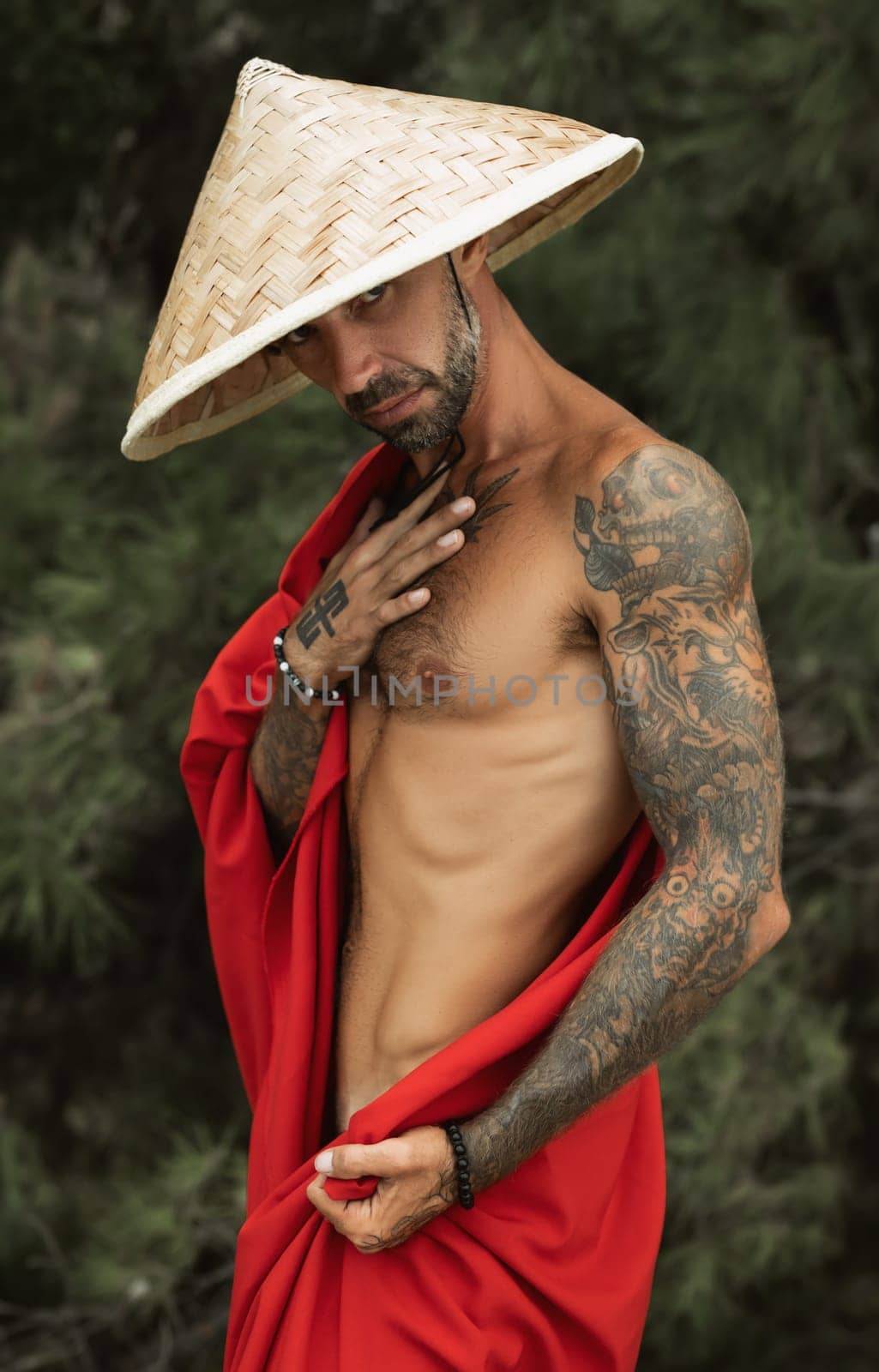 the sexy male monk in a red cape and asian hat in tattoos in nature