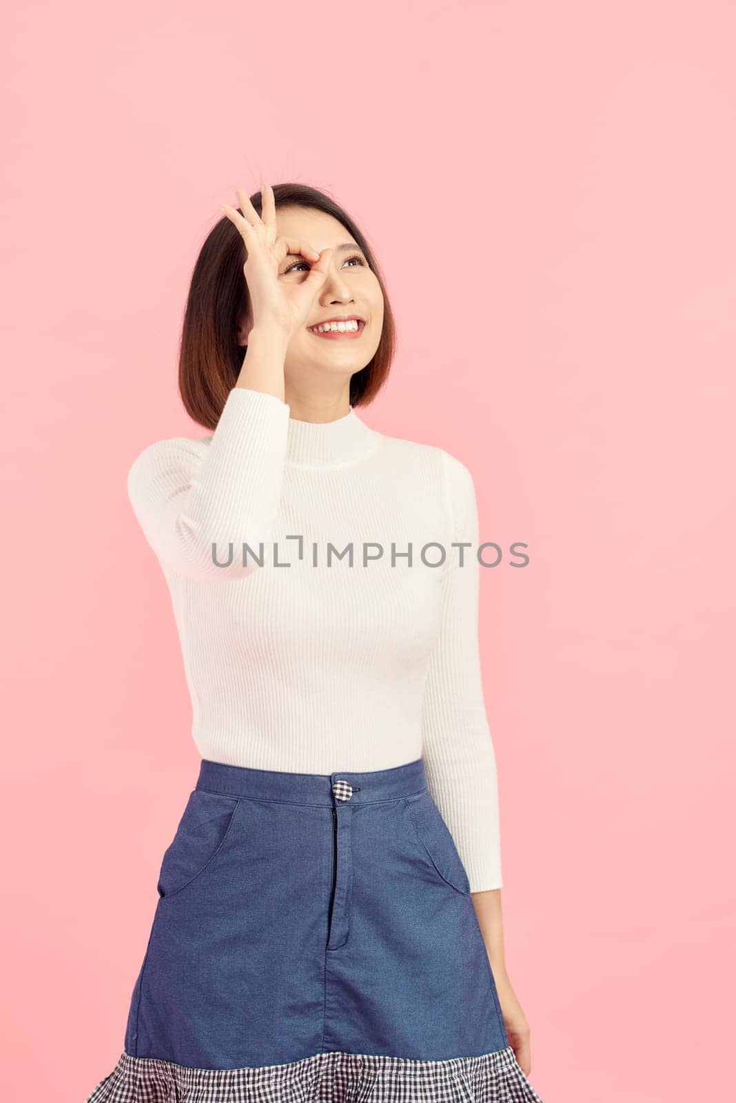 Asian smiling woman showing her hand with ok sign on her eye. Isolated on pink background. by makidotvn
