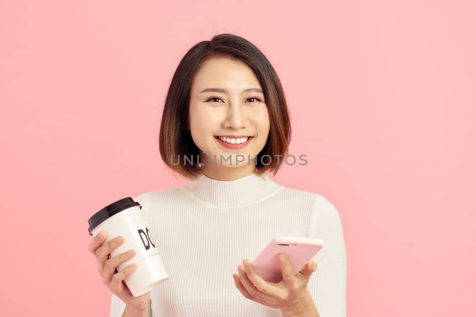 Close up portrait of young Asian woman holding coffee cup and smartphone over pink background.