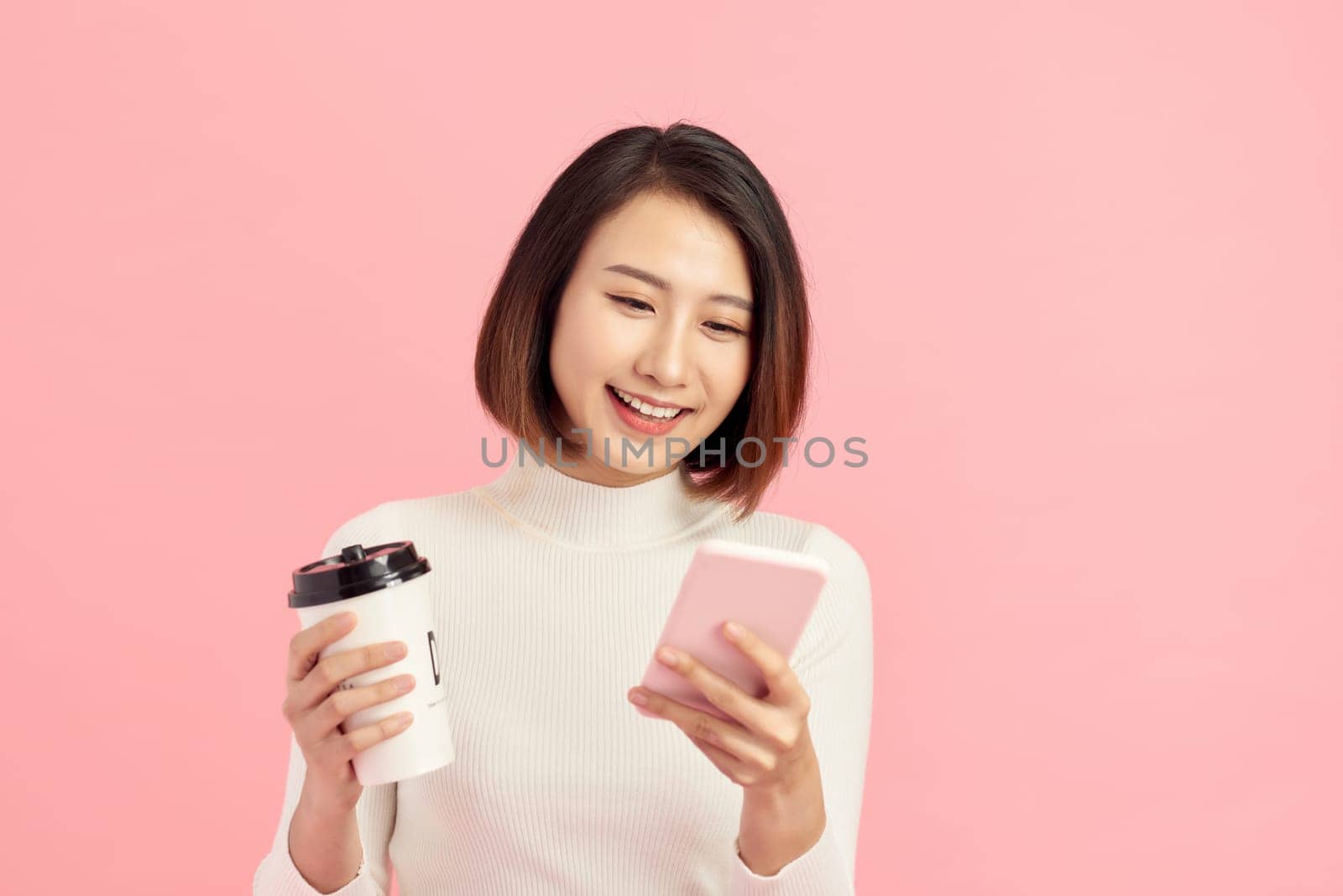 Attractive young Asian woman using mobile phone while holding coffee cup over pink background. by makidotvn