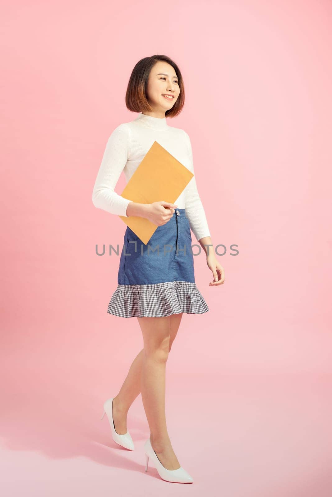 Full length portrait of a happy asian businesswoman holding document while walking isolated over pink background