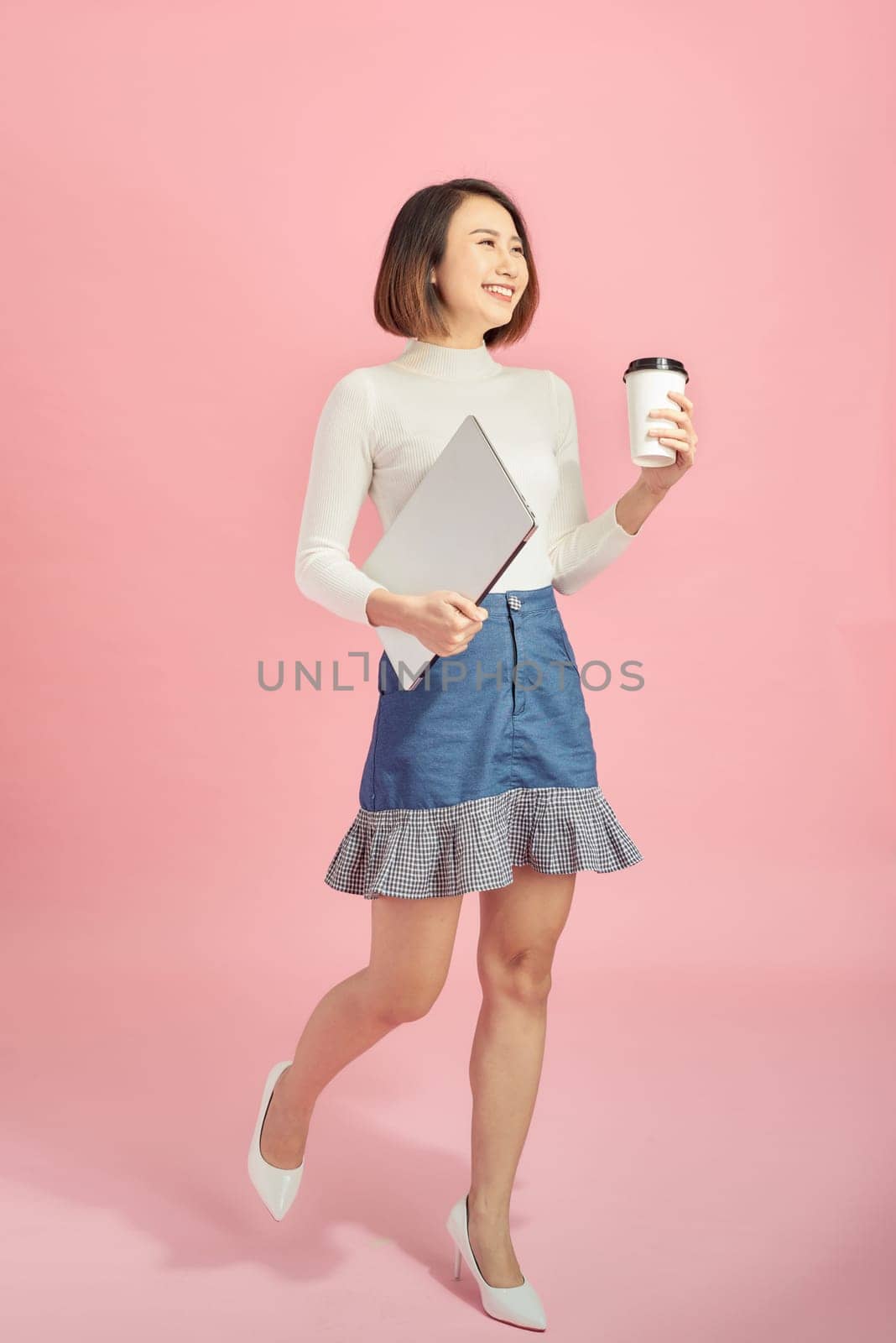 Young Asian woman holding laptop and coffee while walking over pink background.