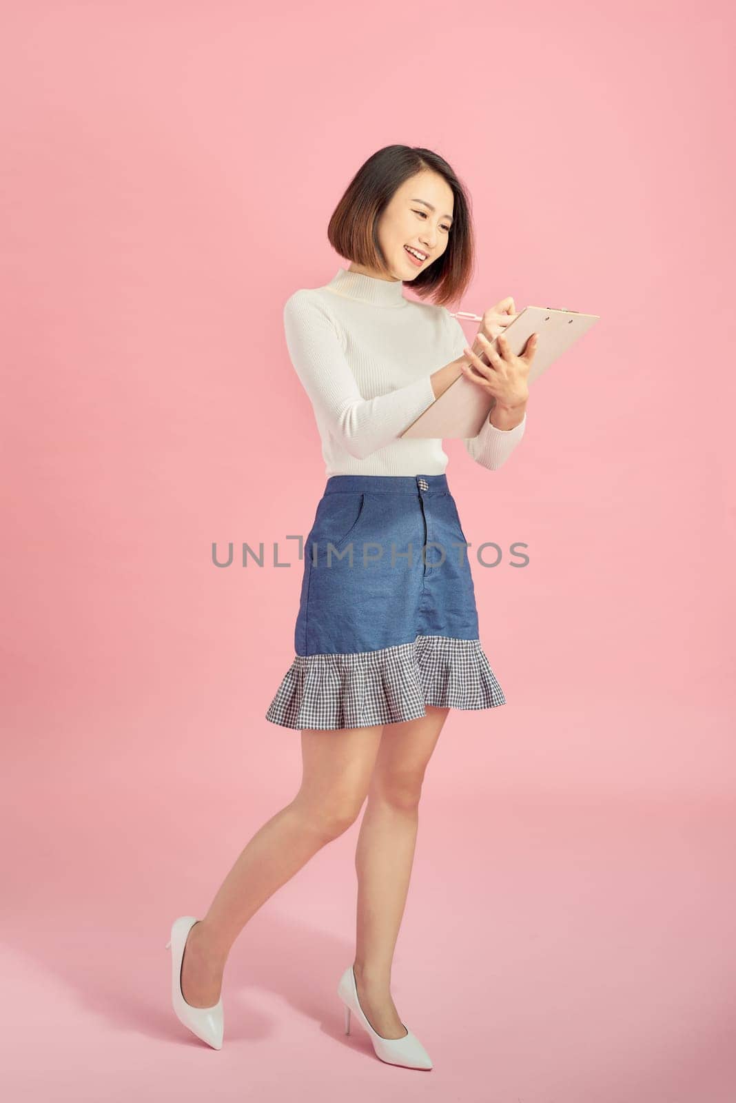 Full length of a smiling Asian woman standing isolated over pink background, writing in the folder. by makidotvn