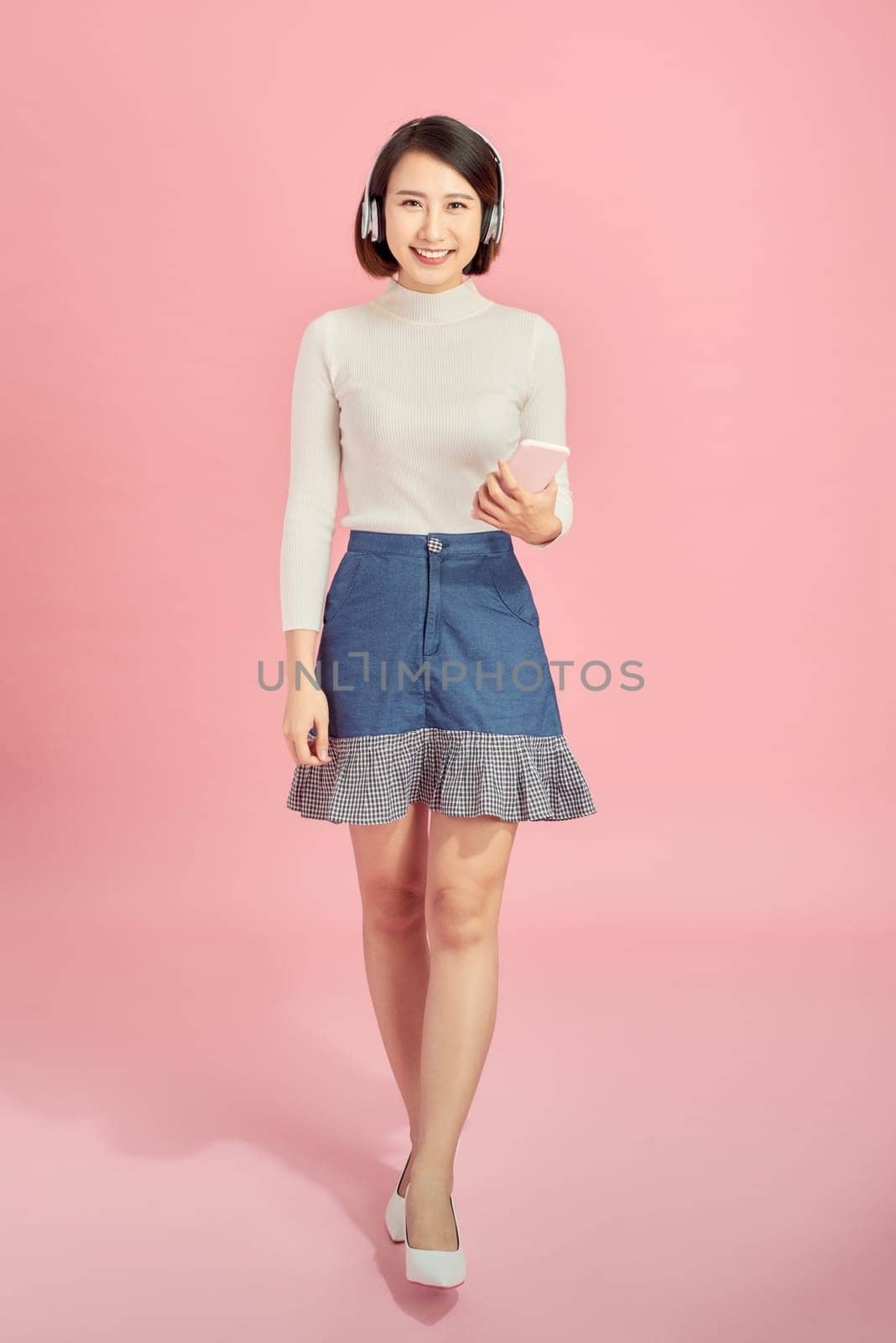 Full length portrait of a happy asian woman with headphones standing and holding mobile phone over pink background