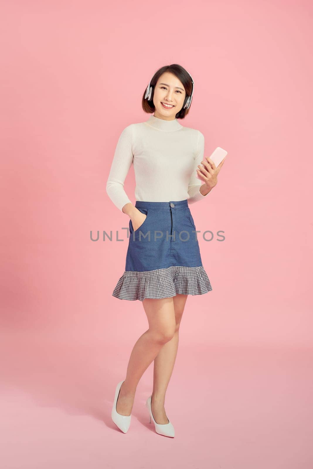 Full length portrait of a happy asian businesswoman listening to music with headphones while dancing isolated over pink background