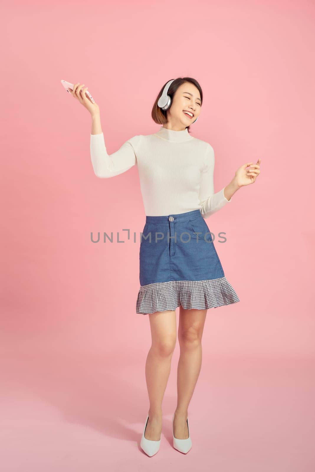 Full length portrait of a happy asian woman with headphones standing and holding mobile phone over pink background by makidotvn