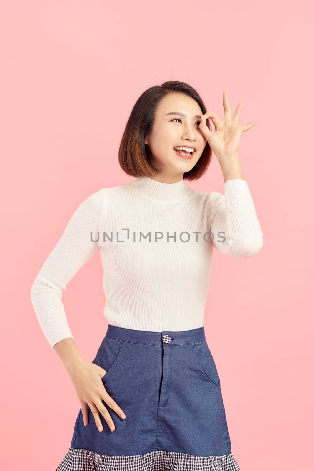 Asian smiling woman showing her hand with ok sign on her eye. Isolated on pink background.