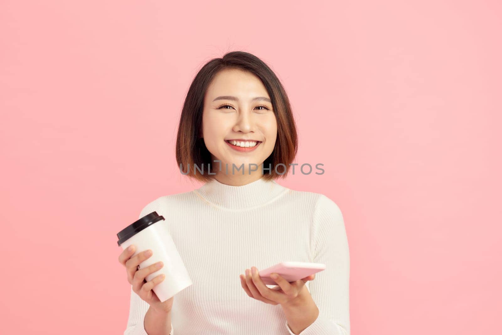 Close up portrait of young Asian woman holding coffee cup and smartphone over pink background. by makidotvn