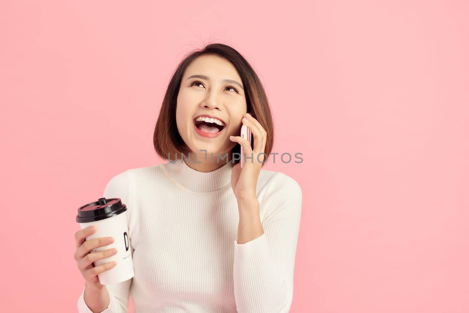 Portrait of cheerful young beautiful Asian woman listening smartphone while holding coffee cup over pink background.