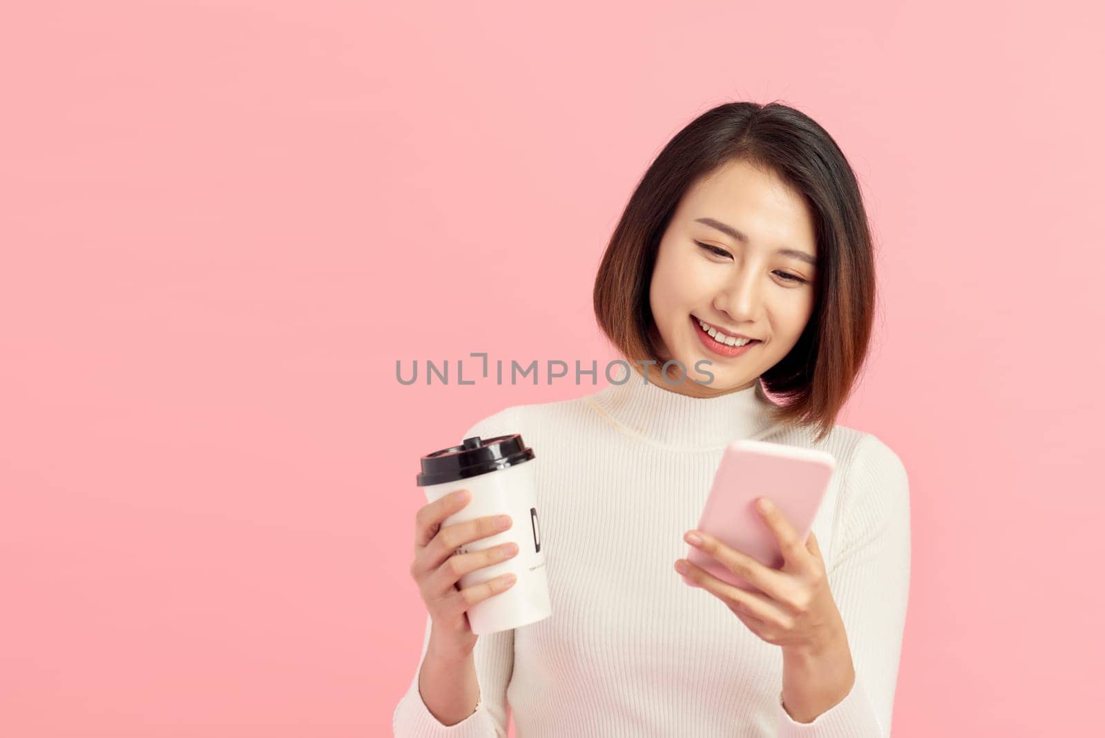 Attractive young Asian woman using mobile phone while holding coffee cup over pink background. by makidotvn