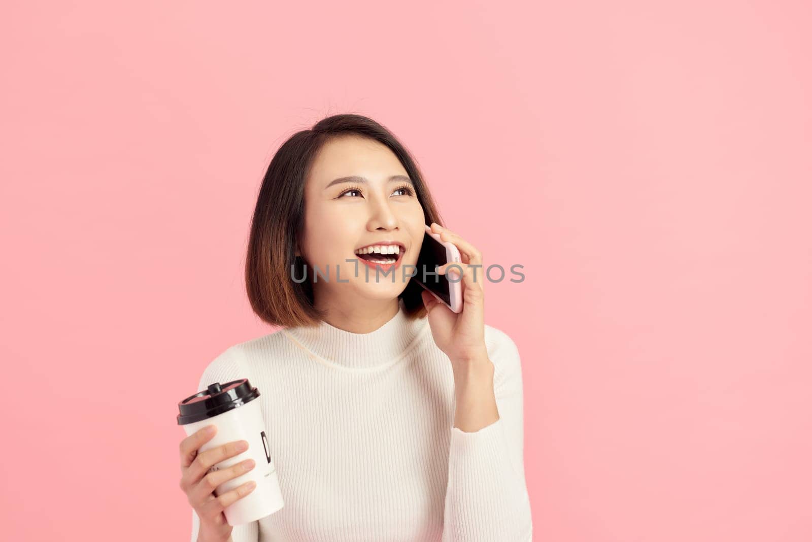 Portrait of cheerful young beautiful Asian woman listening smartphone while holding coffee cup over pink background.