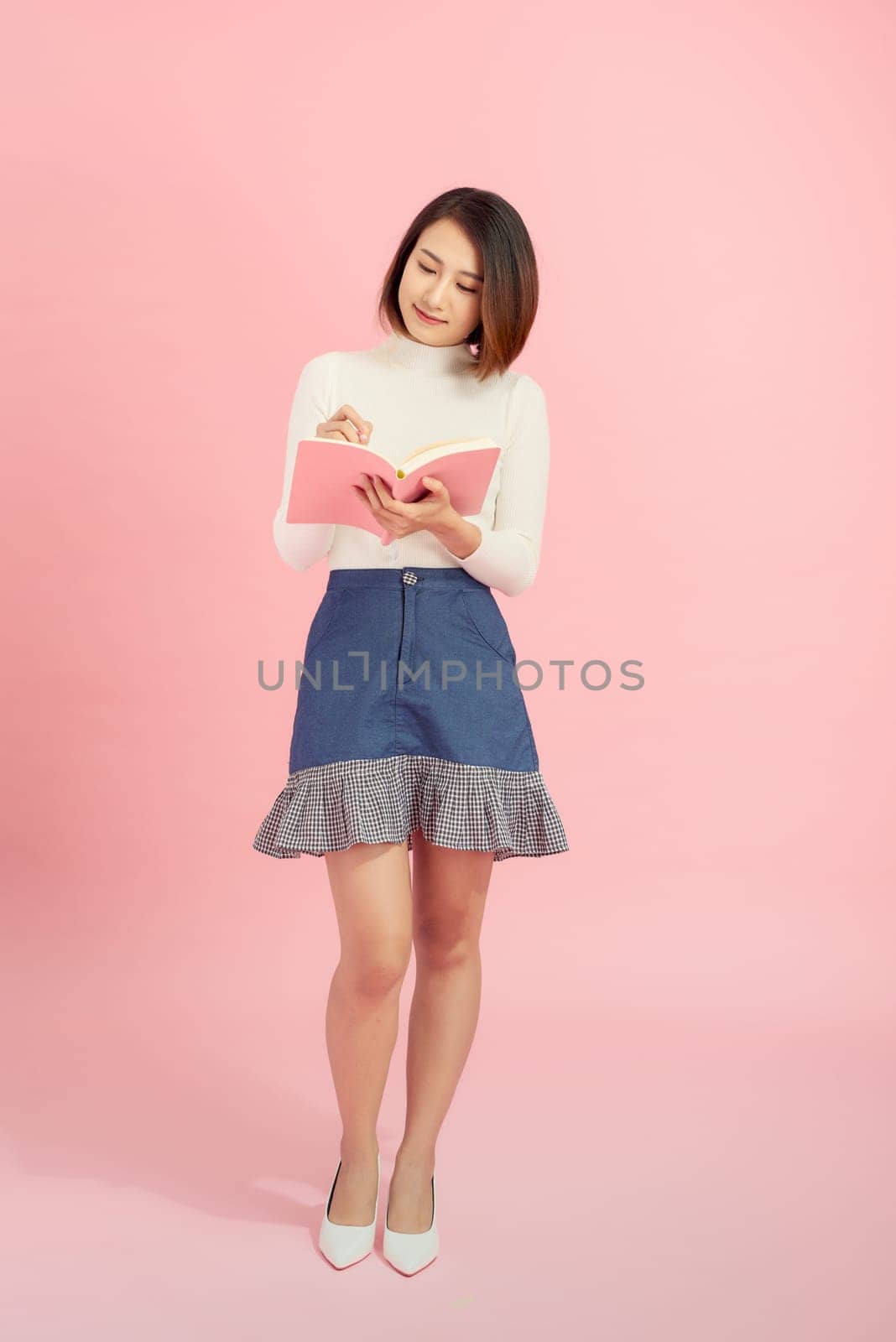 Young asian girl standing, reading interdting book.Isolated on pink background