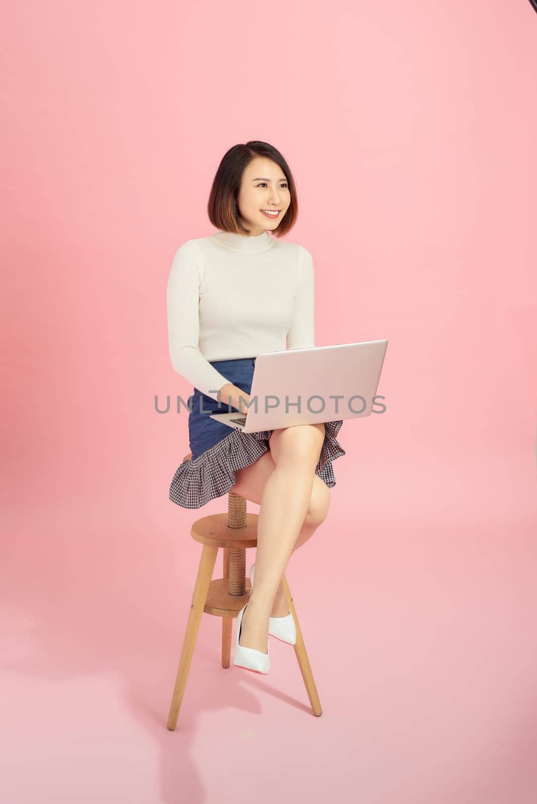 Young beautiful Asian businesswoman using laptop while sitting on chair. Isolated on pink background.