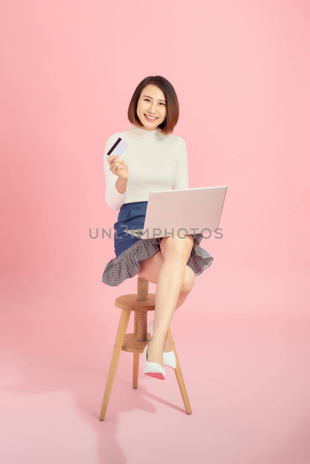 Young Asian woman using credit card to payment online with her laptop. Isolated on pink background. by makidotvn