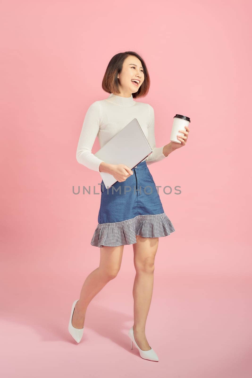 Young Asian woman holding laptop and coffee while walking over pink background. by makidotvn