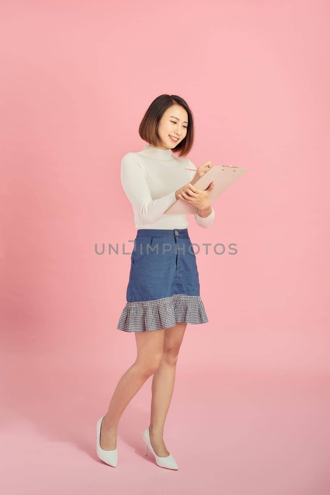 Full length of a smiling Asian woman standing isolated over pink background, holding notepad and pencil by makidotvn