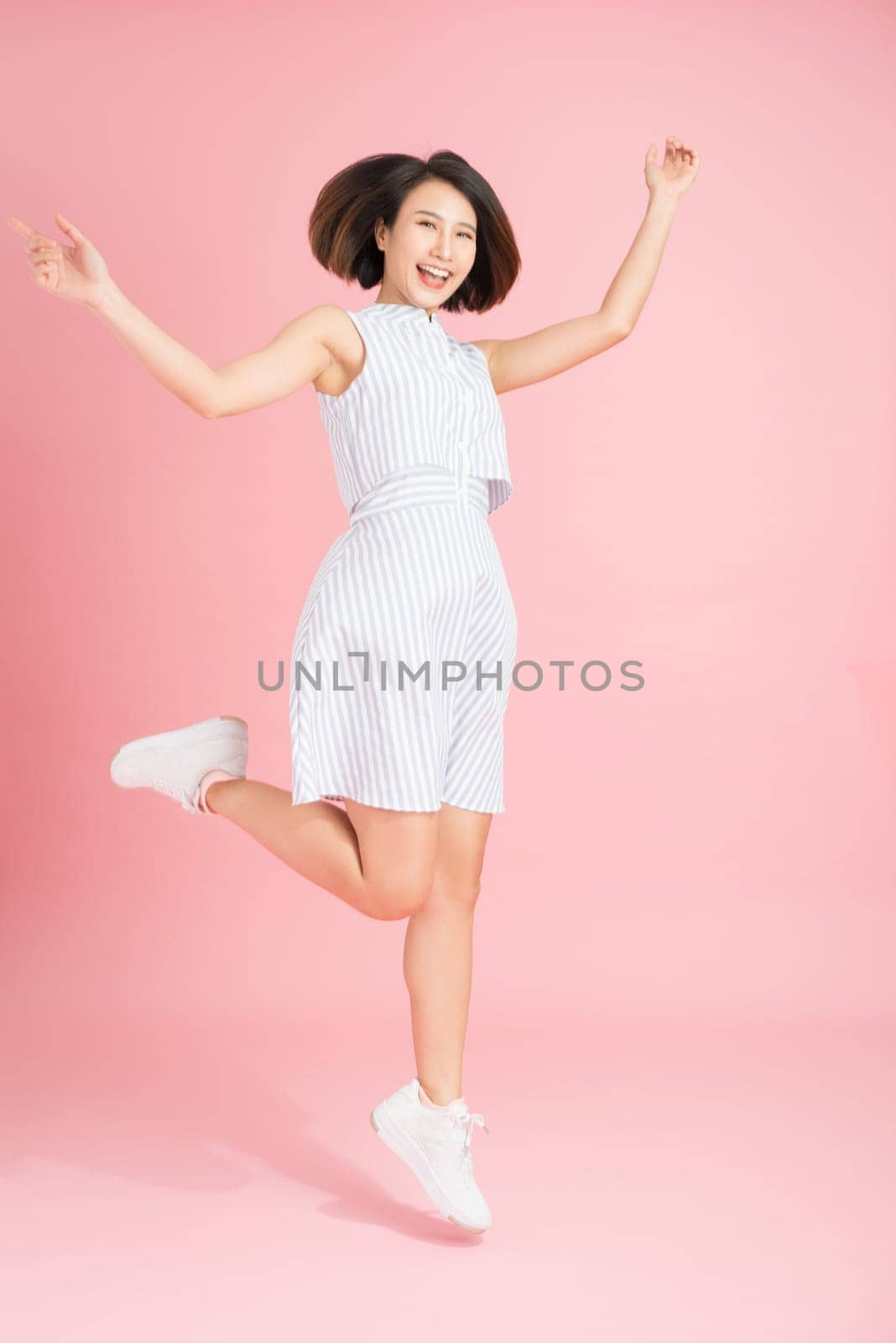 Full length portrait of happy exited pretty girl in elegant light blue dress looking at camera while jumping over pink background by makidotvn