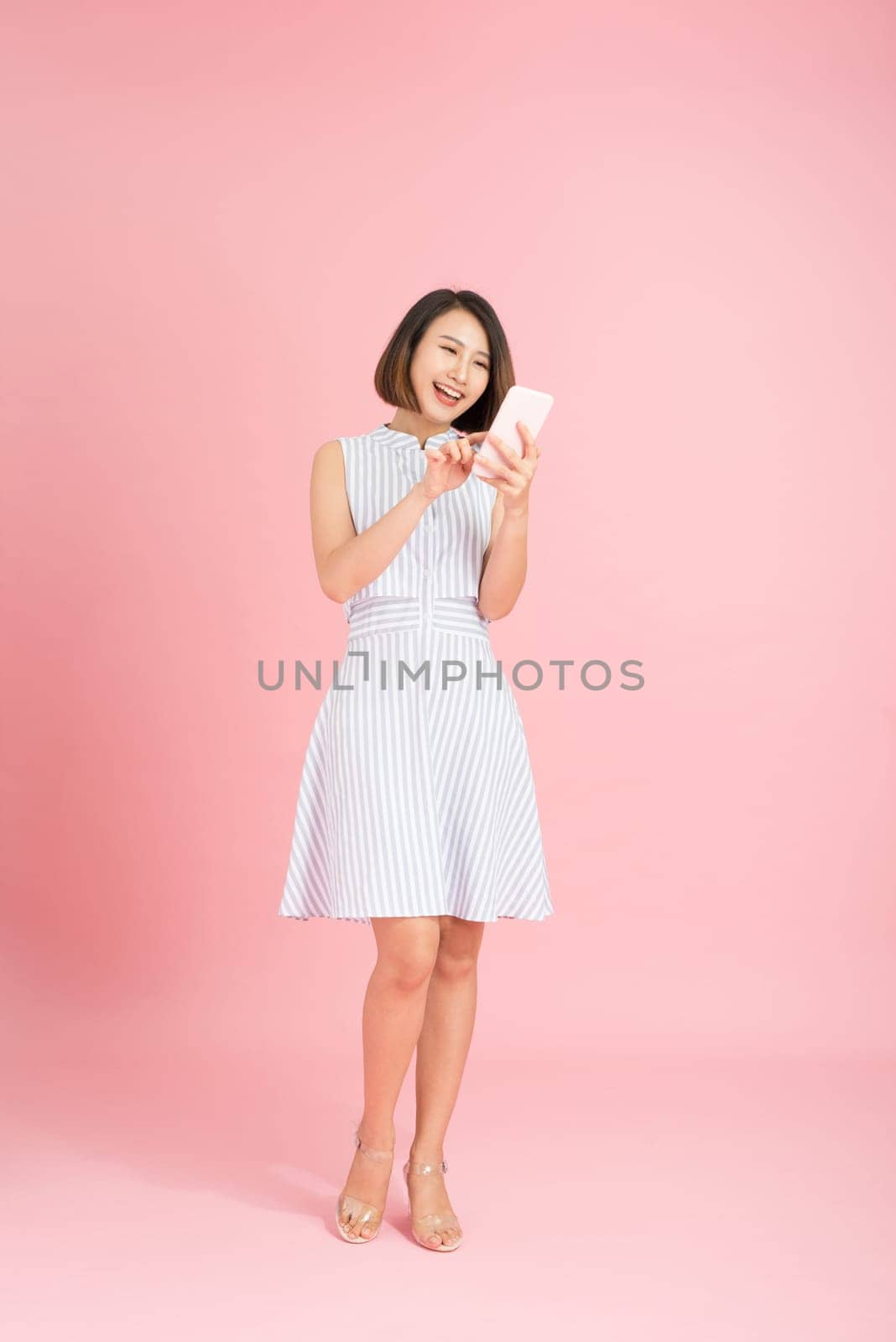 Full length portrait of a smiling casual girl using mobile phone isolated over pink background by makidotvn