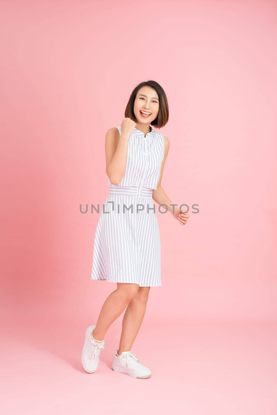 Full length portrait of happy exited pretty girl in elegant light blue dress looking at camera while jumping over pink background by makidotvn