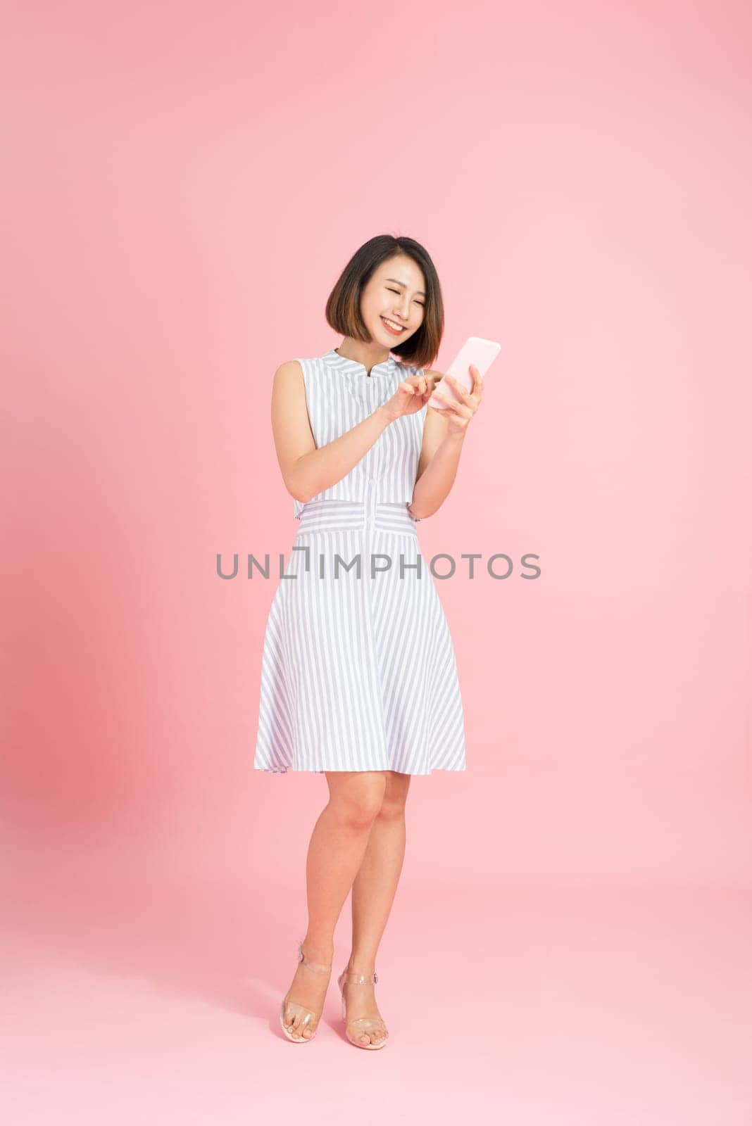Full length portrait of a pretty young woman standing isolated over pink background, using mobile phone