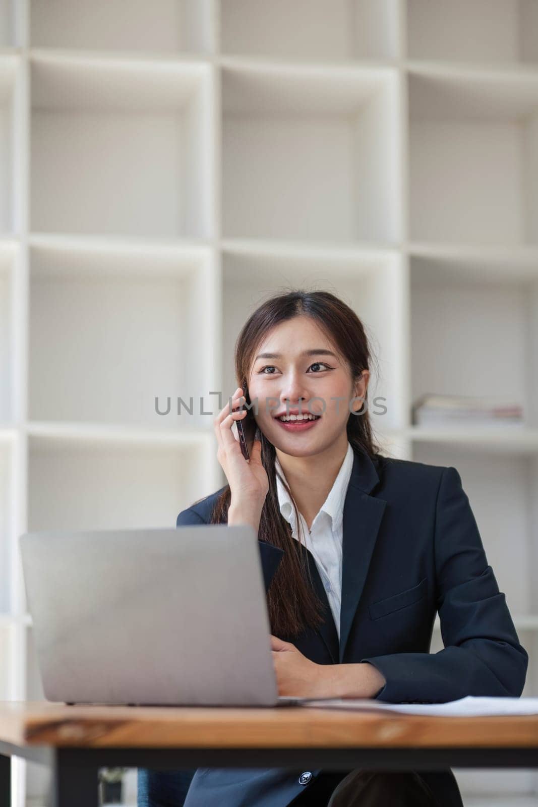 Attractive Asian woman using laptop in online meeting. Businesswoman using mobile phone talking with colleague sitting at table in modern office by wichayada