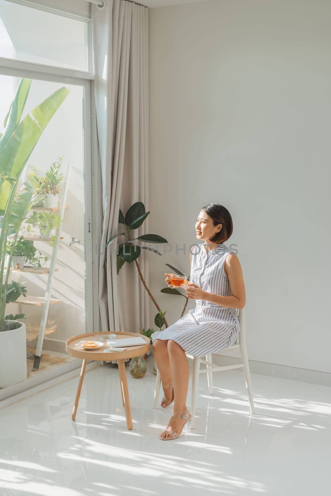 Young charming woman sitting on chair and reading book next to window at home. by makidotvn